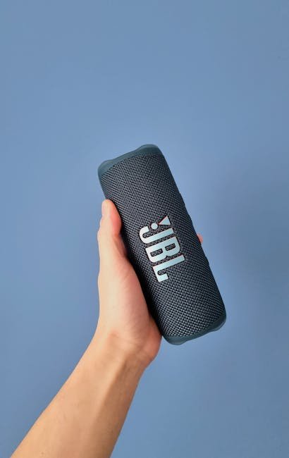 A person's hand holding a sleek portable wireless speaker against a minimalist background.