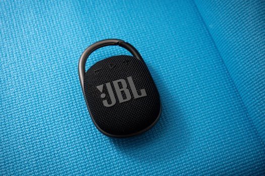 Black JBL portable wireless speaker with carabiner on textured blue background. Ideal for travel and outdoor use.