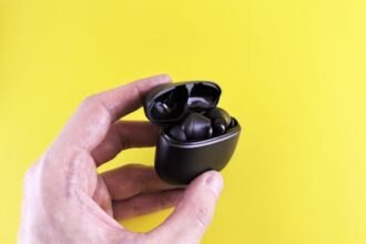 Close-up of a hand holding black wireless Bluetooth earbuds with a yellow backdrop.