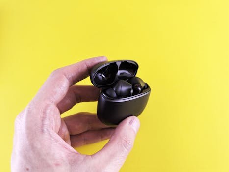 Close-up of a hand holding black wireless Bluetooth earbuds with a yellow backdrop.