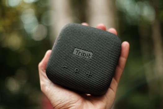 Tibit Stormbox Micro 1 Hand holding a Tribit portable speaker with blurred outdoor background, featuring modern design and textured surface.